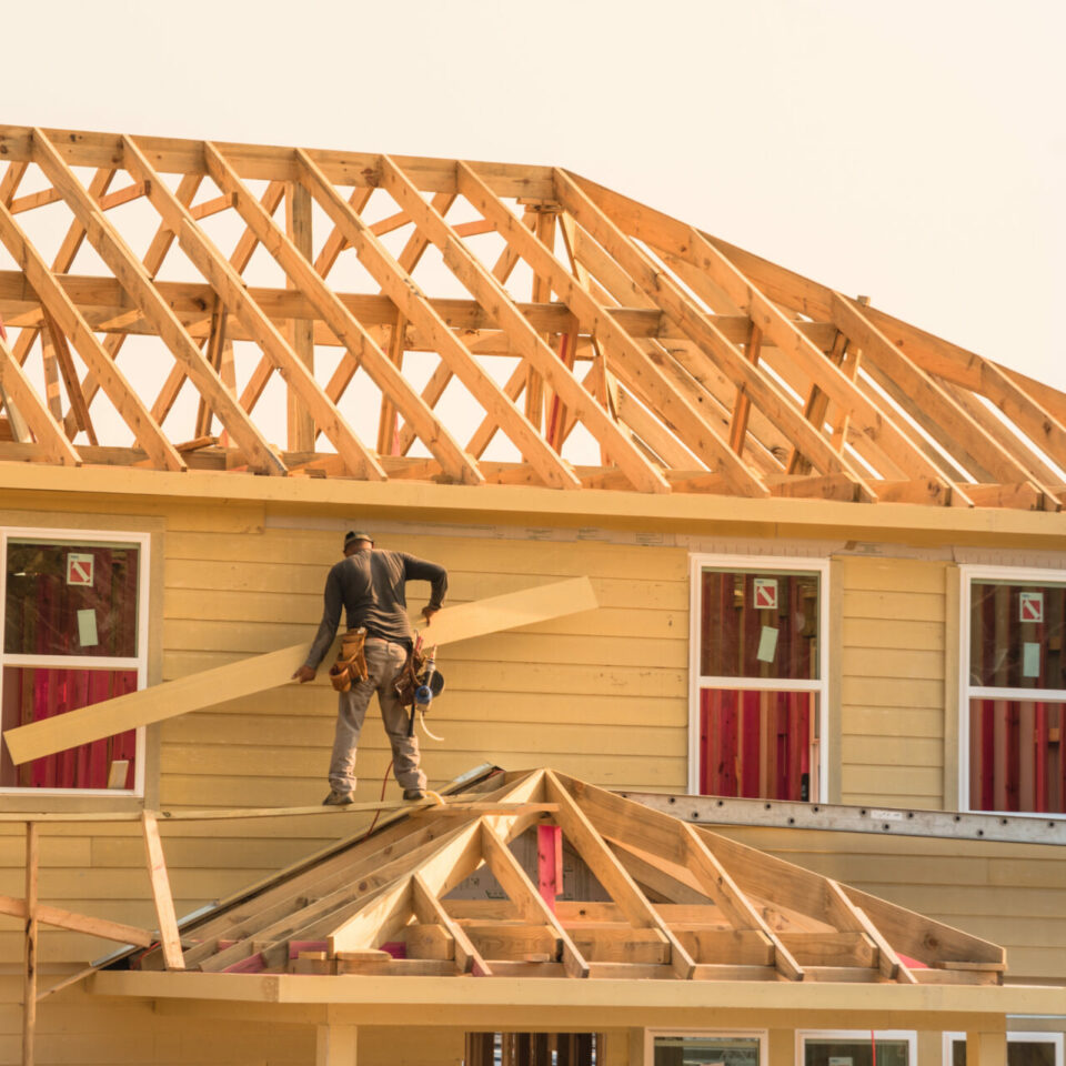 Rear view of builder at eco wooden house construction site near completed suburban home in Humble, Texas, US during sunset. Carpenter on scaffolding installing board of stick built. Labor day concept